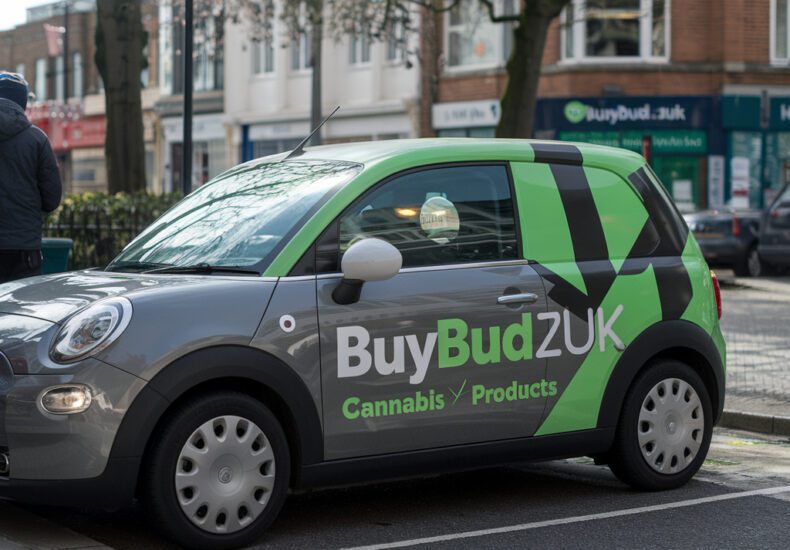 A compact car with BuyBudZUK branding parked on a city street. The vehicle features the company’s green and gray colors with the text "Cannabis Products" on the side, promoting cannabis delivery services in the UK